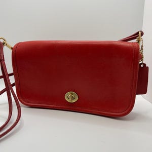May include: A red leather crossbody bag with a gold-tone turn-lock closure. The bag has a long, adjustable strap and a small tag attached to a chain. The bag is rectangular and has a classic, vintage design.