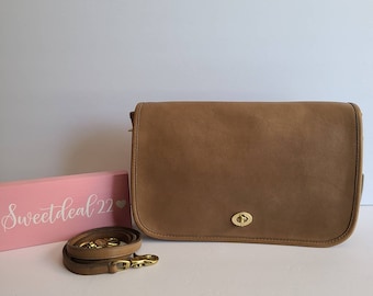May include: A brown leather shoulder bag with a gold clasp and a long adjustable strap. The bag is sitting on a pink surface with the text "Sweetdeal22" written on it.