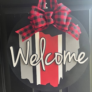 May include: A round wooden door hanger with a black background and a red, white, and gray striped outline of the state of Ohio. The word "Welcome" is written in white cursive letters. A red and black buffalo plaid bow is attached to the top of the hanger.