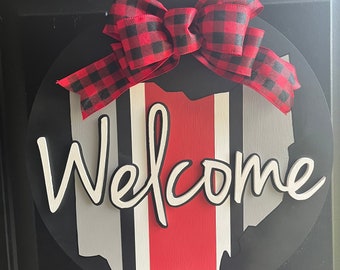 May include: A round wooden door hanger with a black background and a red, white, and gray striped outline of the state of Ohio. The word "Welcome" is written in white cursive letters. A red and black buffalo plaid bow is attached to the top of the hanger.
