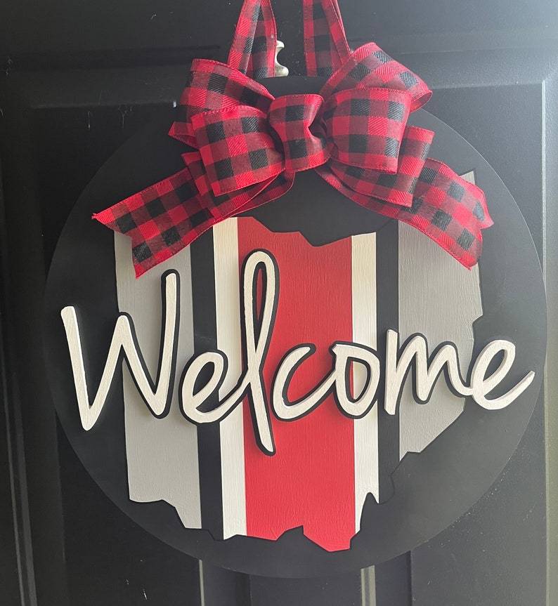 May include: A round wooden door hanger with a black background and a red, white, and gray striped outline of the state of Ohio. The word "Welcome" is written in white cursive letters. A red and black buffalo plaid bow is attached to the top of the hanger.