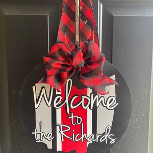 May include: A round wooden door hanger with a black background and a red and white Ohio state outline. The words "Welcome to the Richards" are written in white. A red and black buffalo plaid bow is attached to the top.