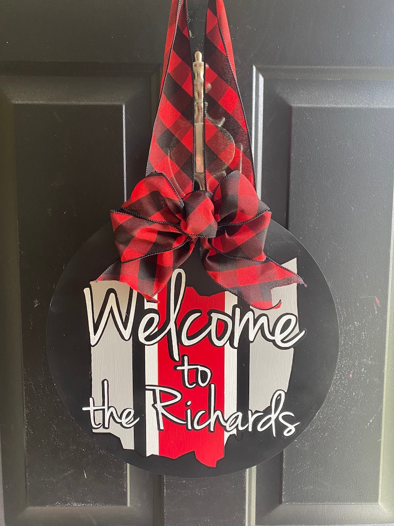 May include: A round wooden door hanger with a black background and a red and white Ohio state outline. The words "Welcome to the Richards" are written in white. A red and black buffalo plaid bow is attached to the top.
