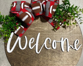 May include: A round wooden welcome sign with a football-themed bow. The sign features the word "Welcome" in white script. The bow has a red background with footballs. Greenery accents the top of the sign.