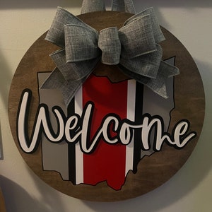 May include: A wooden round door hanger with a gray bow, the outline of the state of Ohio, and the word "Welcome" in white letters.