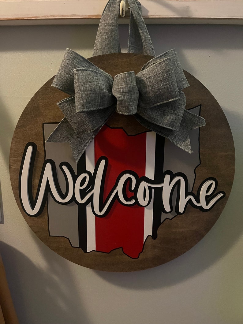 May include: A wooden round door hanger with a gray bow, the outline of the state of Ohio, and the word "Welcome" in white letters.
