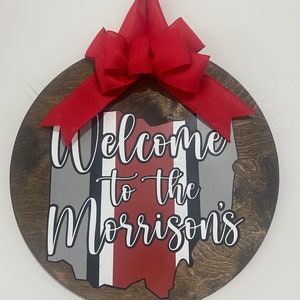 May include: A round wooden door hanger with a red bow. The door hanger features a painted outline of the state of Ohio with the words "Welcome to the Morrisons" in white cursive lettering.