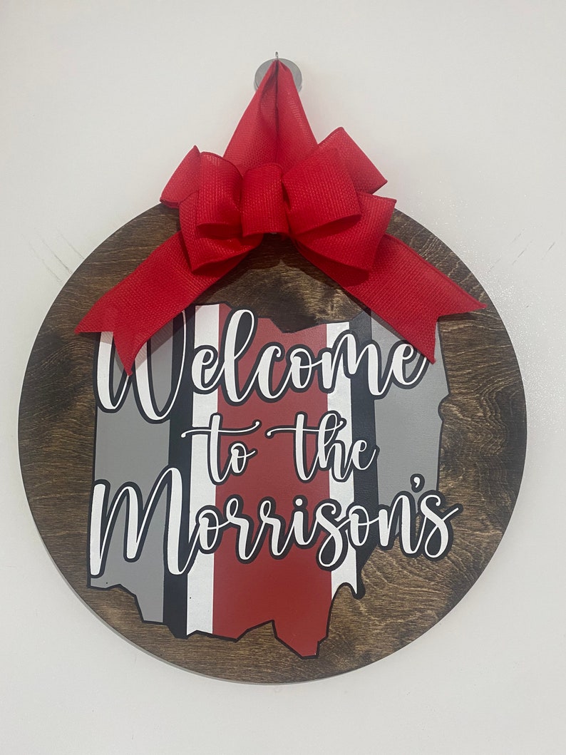 May include: A round wooden door hanger with a red bow. The door hanger features a painted outline of the state of Ohio with the words "Welcome to the Morrisons" in white cursive lettering.