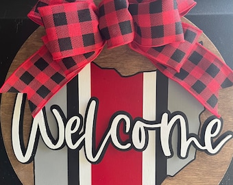 May include: A round wooden door hanger with a red, white, and gray outline of the state of Ohio. The word "Welcome" is written in white with a black outline. A red and black buffalo plaid bow is attached to the top of the door hanger.