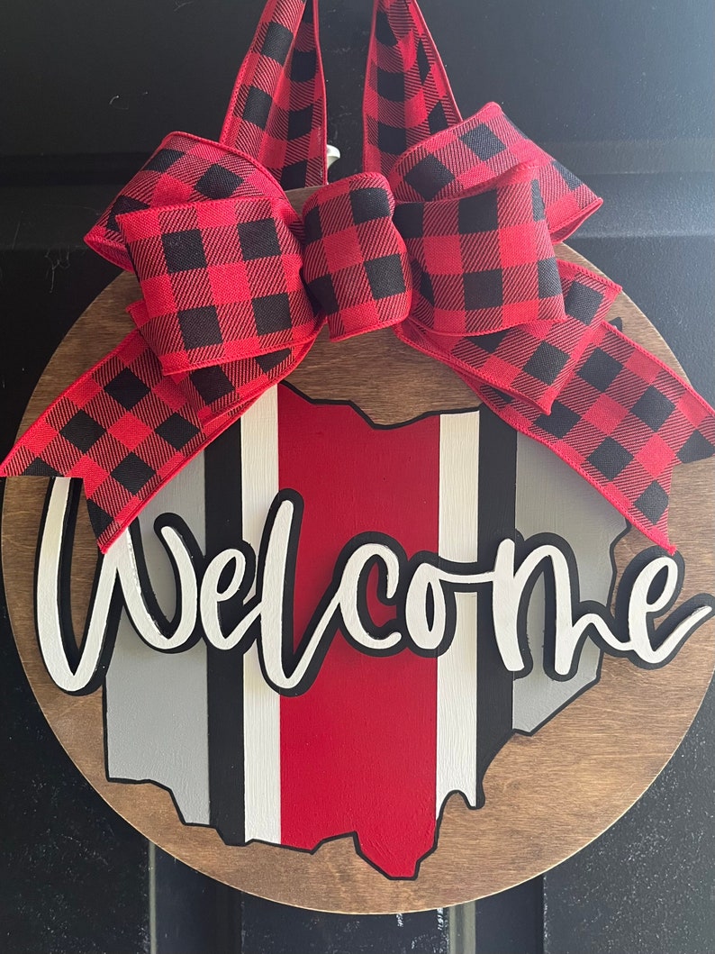 May include: A round wooden door hanger with a red, white, and gray outline of the state of Ohio. The word "Welcome" is written in white with a black outline. A red and black buffalo plaid bow is attached to the top of the door hanger.