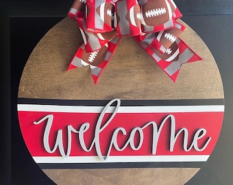 May include: A round wooden door hanger with a red, white, and black striped band and the word "welcome" in white lettering. A red and gray bow with a football pattern is attached to the top.