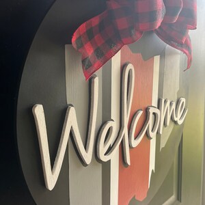 May include: A round wooden welcome sign with a black background and white lettering. The sign has vertical stripes in red, white, and gray. A red and black plaid bow is attached to the top of the sign.