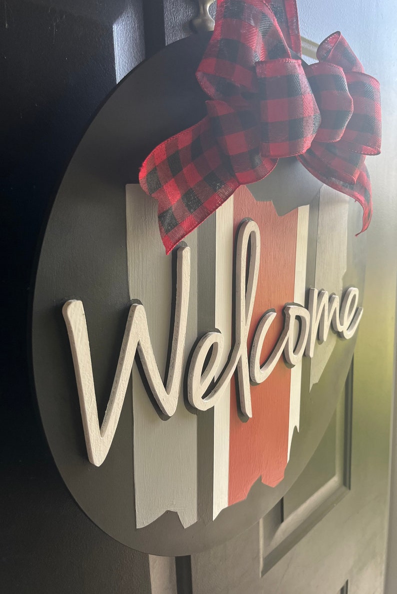 May include: A round wooden welcome sign with a black background and white lettering. The sign has vertical stripes in red, white, and gray. A red and black plaid bow is attached to the top of the sign.