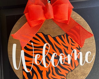 May include: A round wooden door hanger with an orange and black tiger stripe design in the shape of the state of Ohio. The words "Welcome" are written in white cursive lettering. A large orange bow is attached to the top of the hanger.