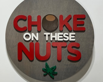 May include: A round wooden sign with the phrase "CHOKE ON THESE NUTS" in red and white letters. A brown buckeye and a green leaf are also on the sign. The sign is gray with a brown leather strap for hanging.