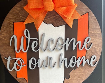 May include: A wooden round door hanger with an orange, brown, and white striped outline of the state of Ohio. The words "Welcome to our home" are written in a silver cursive font. An orange bow is attached to the top of the hanger.