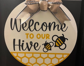 May include: A round wooden door hanger with a white background and a yellow honeycomb pattern. The text "Welcome to our Hive" is written in black cursive lettering. Two black and yellow bees are on the right side of the sign. A brown and black checkered bow is at the top of the sign.