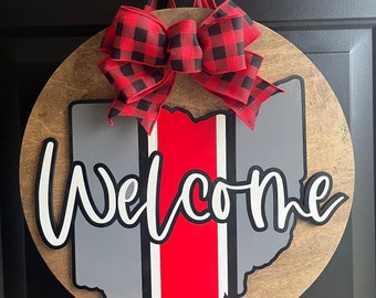 May include: A round wooden door hanger with a red, white, and gray outline of the state of Ohio. The word "Welcome" is written in white cursive lettering. A red and black buffalo plaid bow is attached to the top of the hanger.