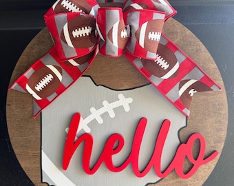 May include: A round wooden door hanger featuring a gray Ohio outline with a white football design and the red word "hello." A large bow with football-themed ribbon tops the sign, perfect for football season.