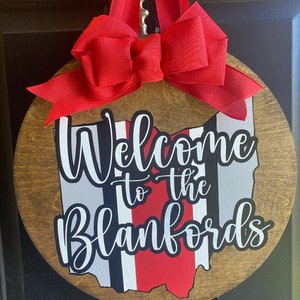 May include: A round wooden door hanger with a red bow. The door hanger features a white outline of the state of Ohio with red and black stripes. The text "Welcome to the Blankfords" is written in white cursive lettering.