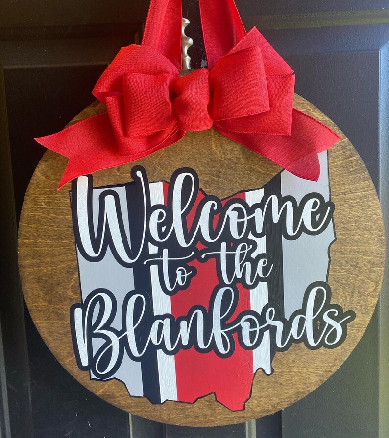 May include: A round wooden door hanger with a red bow. The door hanger features a white outline of the state of Ohio with red and black stripes. The text "Welcome to the Blankfords" is written in white cursive lettering.