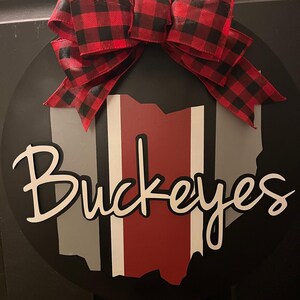May include: A black and white round wooden sign with a red, white, and gray outline of the state of Ohio. The sign says "Buckeyes" in white cursive lettering. A red and black buffalo plaid bow is attached to the top of the sign.