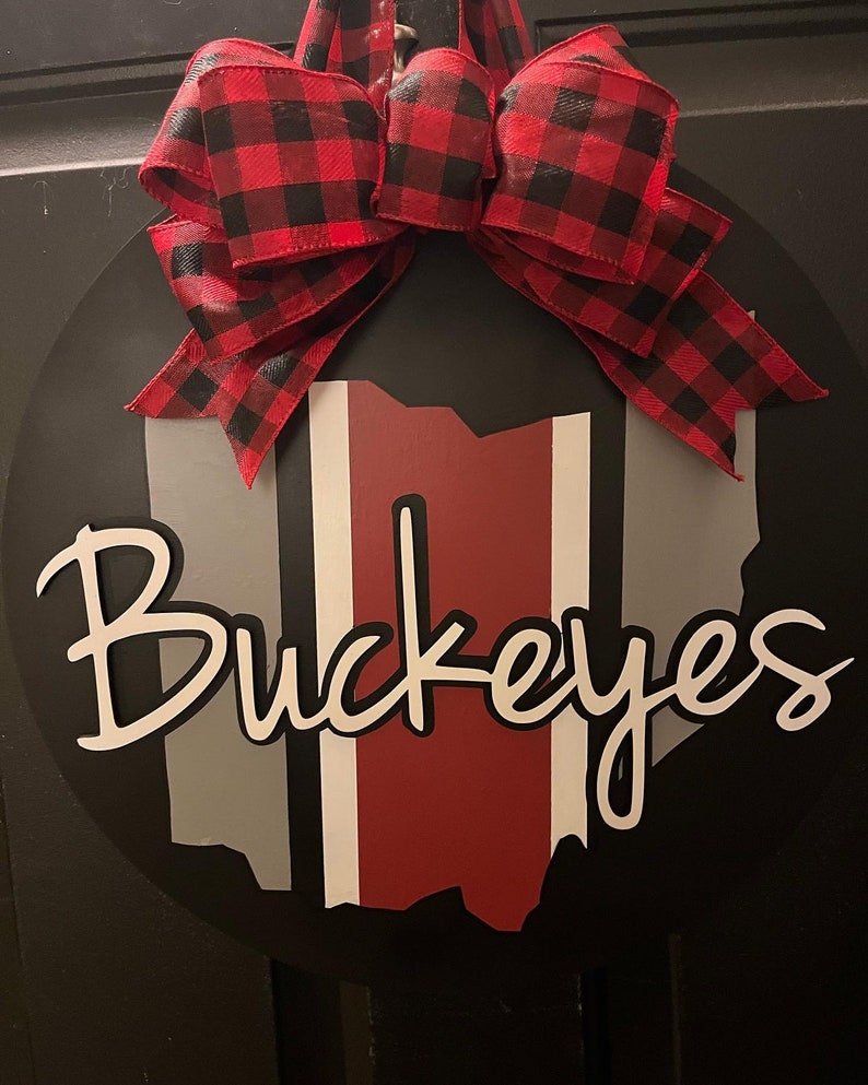 May include: A black and white round wooden sign with a red, white, and gray outline of the state of Ohio. The sign says "Buckeyes" in white cursive lettering. A red and black buffalo plaid bow is attached to the top of the sign.
