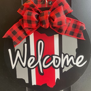 May include: A black and red welcome sign with a red and black buffalo plaid bow. The sign features a white outline of the state of Ohio with red and white stripes.