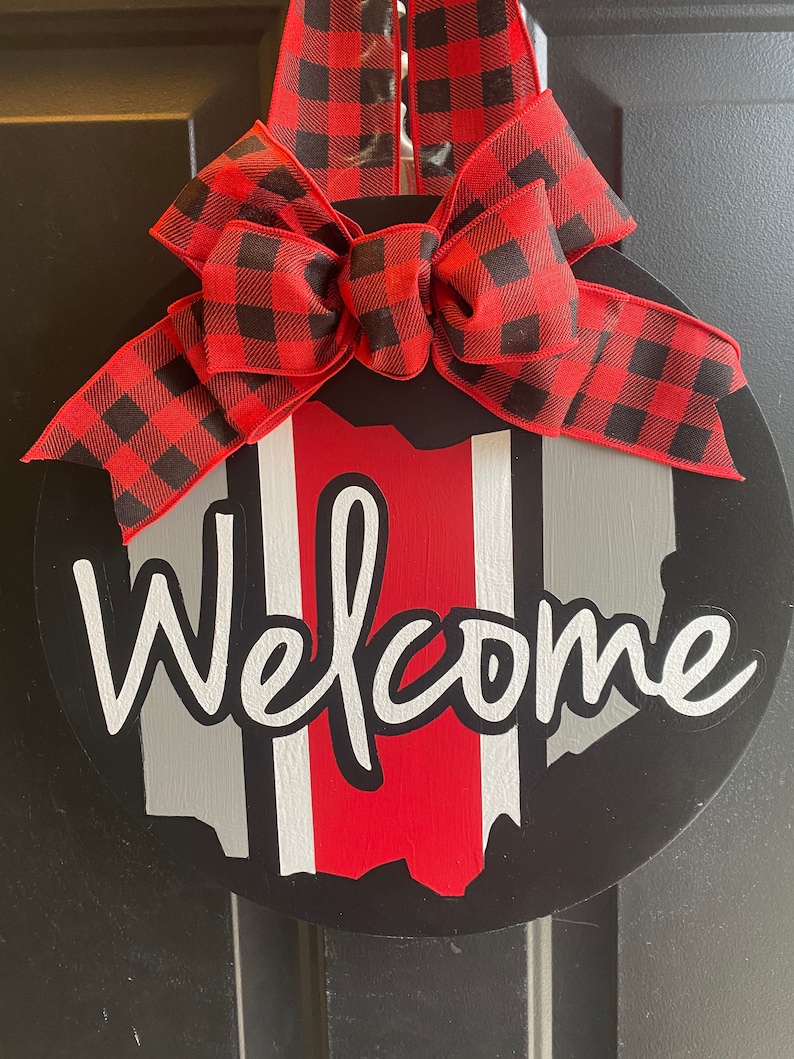 May include: A black and red welcome sign with a red and black buffalo plaid bow. The sign features a white outline of the state of Ohio with red and white stripes.
