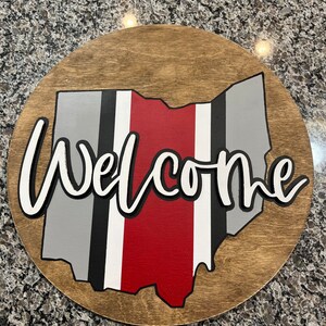 May include: A round wooden sign with the state of Ohio painted in gray, black, white, and red. The word "Welcome" is written in white with a black outline.