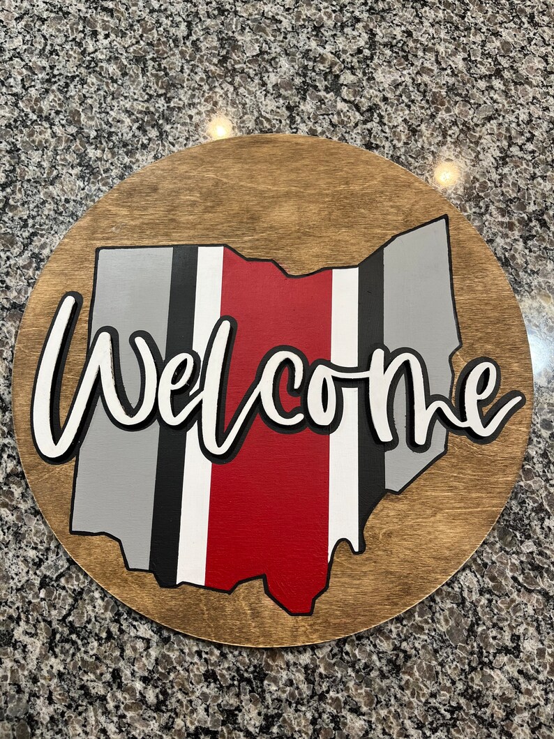May include: A round wooden sign with the state of Ohio painted in gray, black, white, and red. The word "Welcome" is written in white with a black outline.