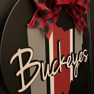 May include: A black and white wooden sign with a red, white, and gray Ohio state outline and the word "Buckeyes" in white script. The sign is decorated with a red and black plaid bow.