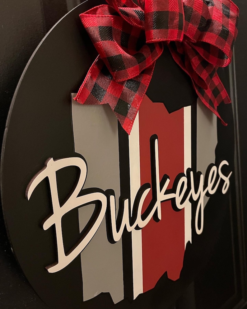 May include: A black and white wooden sign with a red, white, and gray Ohio state outline and the word "Buckeyes" in white script. The sign is decorated with a red and black plaid bow.