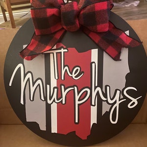 May include: A black and white round wooden sign with a red, white, and gray outline of the state of Ohio. The sign has a red and black buffalo plaid bow on top and the text "The Murphys" in white cursive lettering.