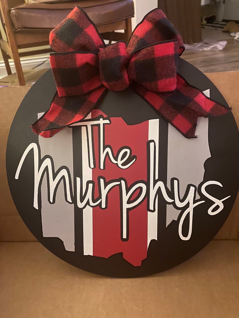 May include: A black and white round wooden sign with a red, white, and gray outline of the state of Ohio. The sign has a red and black buffalo plaid bow on top and the text "The Murphys" in white cursive lettering.
