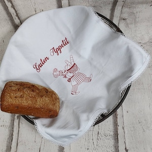 May include: White linen tea towel with red embroidery of a person carrying a basket of bread and the text "Guten Appetit". A loaf of bread is sitting on the towel.