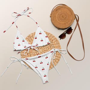 May include: A white bikini with a cherry print. The bikini top is a triangle style with adjustable straps. The bikini bottom is a high-cut style with ties on the sides. The bikini is laying on a woven straw mat.