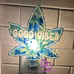 May include: A white night light shaped like a marijuana leaf with a blue and green watercolor pattern. The leaf has the words "GOOD VIBES" printed on it in white.