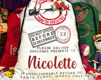 Pode incluir: Um saco de presente de tecido cor de creme com um carimbo vermelho do Polo Norte com a silhueta de um trenó, texto preto DO NOT OPEN BEFORE 25 DECEMBER e o nome Nicolette em glitter vermelho. Inclui cordões pretos na parte superior.