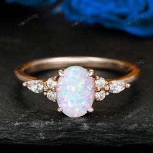 May include: Rose gold ring featuring an oval opal center stone and accent diamonds. The ring has a delicate band and is set against a dark background with blue floral accents. A beautiful piece of jewelry.