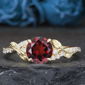 May include: A gold ring featuring a round, deep red garnet gemstone at the center. The band is designed with intricate metal leaf motifs and small, clear, round-cut accent stones embedded along the sides of the shank, creating a nature-inspired look.