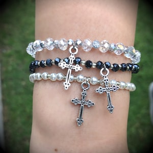 May include: Three layered bracelets with silver cross charms. The top bracelet is made of clear faceted beads, the middle bracelet is made of black beads, and the bottom bracelet is made of white beads.