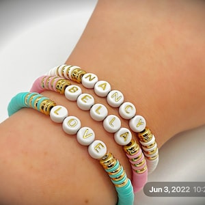 May include: Three colorful bracelets with white letter beads spelling "LOVE", "BELLA", and "NANCY". The bracelets feature gold accents and are in shades of pink, turquoise, and white. The bracelets are made of flat, disc-shaped beads.