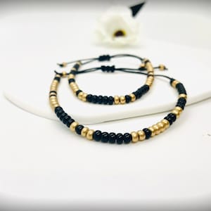 Puede incluir: Dos pulseras a juego con cuentas negras y doradas. Las pulseras están sobre una superficie blanca. Las cuentas están ensartadas en un cordón negro y las pulseras tienen cierres ajustables. Un accesorio elegante.