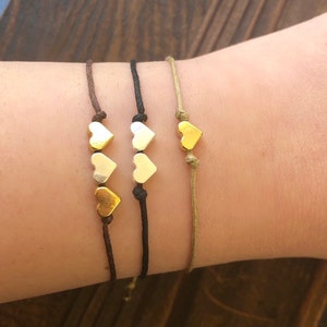 Könnte beinhalten: Drei Freundschaftsarmbänder mit goldenen Herzanhängern. Die Armbänder sind aus braunem, schwarzem und beigefarbenem Band gefertigt.
