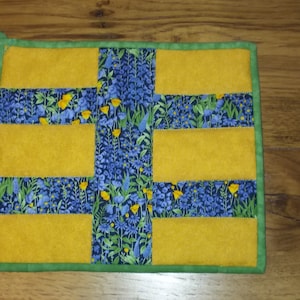 May include: A rectangular pot holder with a floral pattern and yellow accents. The center features a blue floral fabric with yellow flowers. Yellow rectangles are sewn on top. The edges are trimmed with green fabric and a green loop.