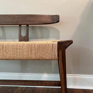 May include: A wooden bench with a curved back and a woven seat. The bench is made of dark wood and has a natural finish. The seat is made of a light brown woven material.