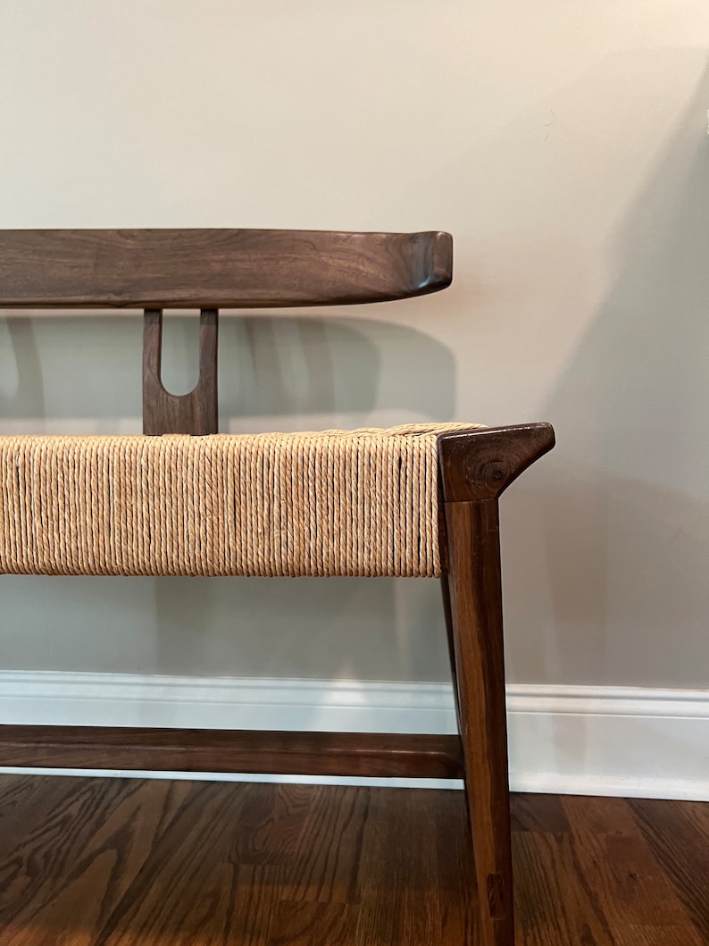 May include: A wooden bench with a curved back and a woven seat. The bench is made of dark wood and has a natural finish. The seat is made of a light brown woven material.