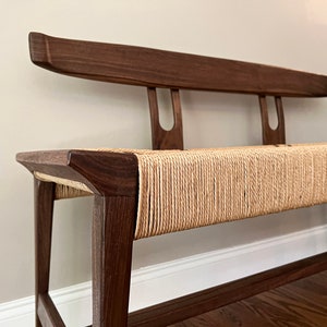 May include: A wooden bench with a natural woven rope seat and a dark brown wood frame. The bench has a simple, modern design with a slightly curved backrest.