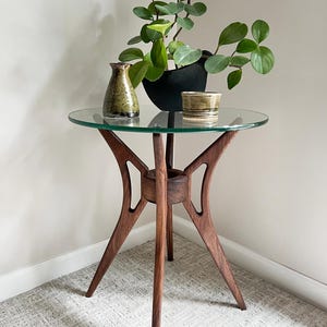 May include: A mid-century modern style side table with a round glass top and a dark wood base. The table supports a potted plant, a small vase, and a decorative bowl. The table is approximately 24 inches in height.